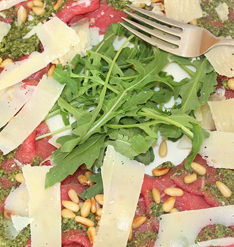 Picture of Beef Carpaccio with flakes of parmesan cheese on top of it and some salad and  pine nuts